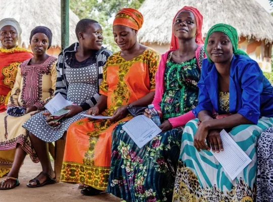 A photo of a group of African women in traditional dress, sat in a row on a bench holding paperwork while they wait for appointments.