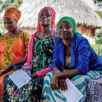 A photo of a group of African women in traditional dress, sat in a row on a bench holding paperwork while they wait for appointments.