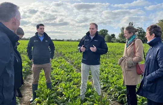 Photo of a group of six business people in a field of crops. One man is talking whilst the others listen.