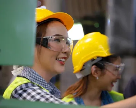 A photo of two smiling female factory workers in hard hats and protective goggles.