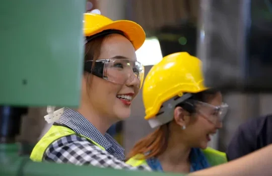 A photo of two smiling female factory workers in hard hats and protective goggles.