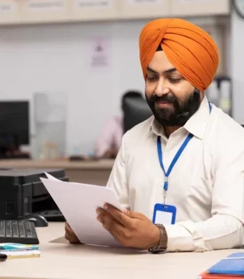 A photo of an Asian man with an orange turban working in an office, looking at paperwork.