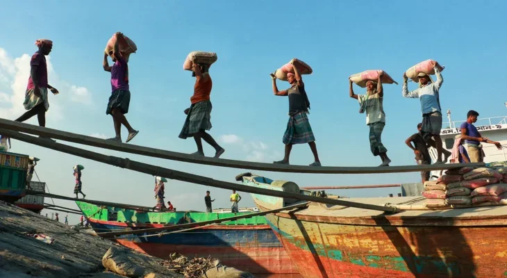 A photo of a group of Asian workers walking along a plant from a boat to the shore, carrying sacks on their heads.