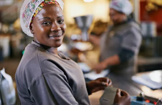 A photo of a smiling black woman wearing a hair net, working in a food factory.