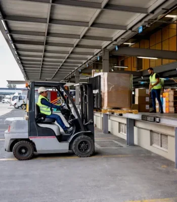 A photo of a man driving a fork lift truck at a factory loading bay.