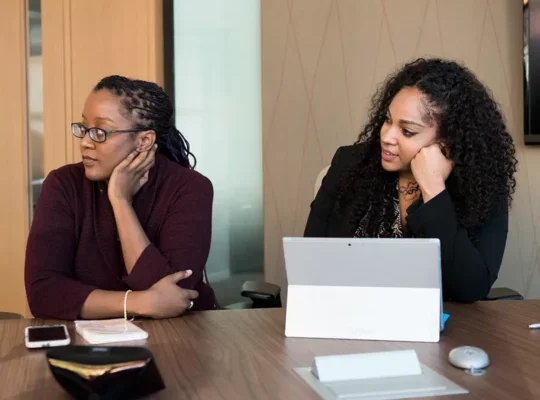 Two women in a meeting room listening to someone speak