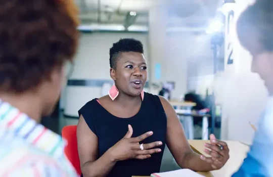 A photo of a middle-aged professional black woman talking to others in an office.