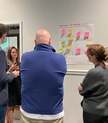 A photo of a group of AIM-Progress members having a discussion by a wall with Post-It notes on it.