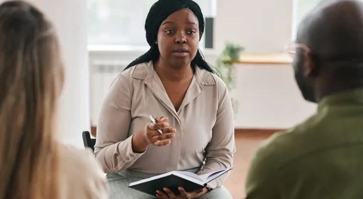 Photo of a woman holding a notebook, speaking to two people in a meeting room.