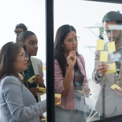 Group of colleagues collaborating in an office, looking pensively at some sticky notes on a glass wall.